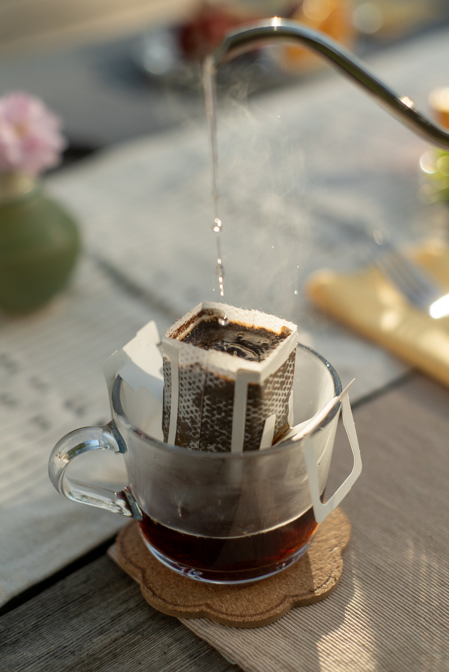 A glass mug with a Kefi Coffee pour-over coffee filter attached is placed on a wooden table. Hot water is being poured from a gooseneck kettle into the filter, with steam rising from the coffee. The mug rests on a cork coaster, and the background shows a blurred dining setup with utensils and a vase with a flower, creating a cozy and inviting atmosphere, like an outdoor picnic.