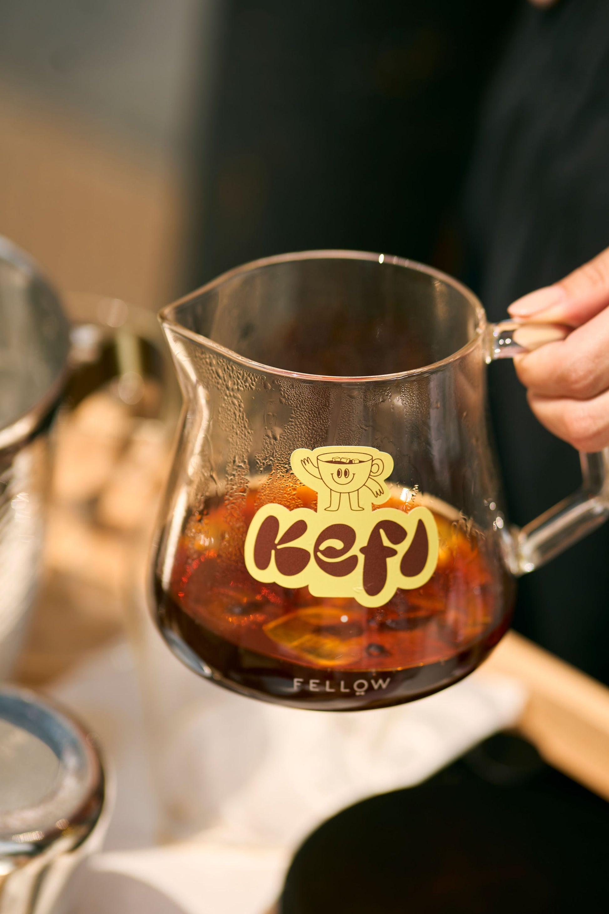 A hand holds a glass carafe featuring the Kefi Coffee logo, a playful character enjoying coffee, filled with freshly brewed dark coffee. The carafe rests on a table, with a cozy, warm ambiance surrounding the brewing setup. The rich, dark coffee inside contrasts with the clear glass and the yellow logo, drawing focus to the coffee and branding. Other brewing tools are softly blurred in the background, adding to the inviting atmosphere.