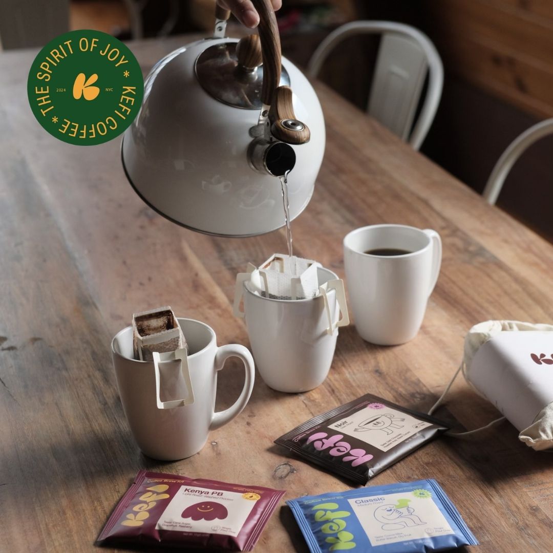 Water being poured from a kettle into cups on a wooden table with Kefi Coffee bags and packaging.
