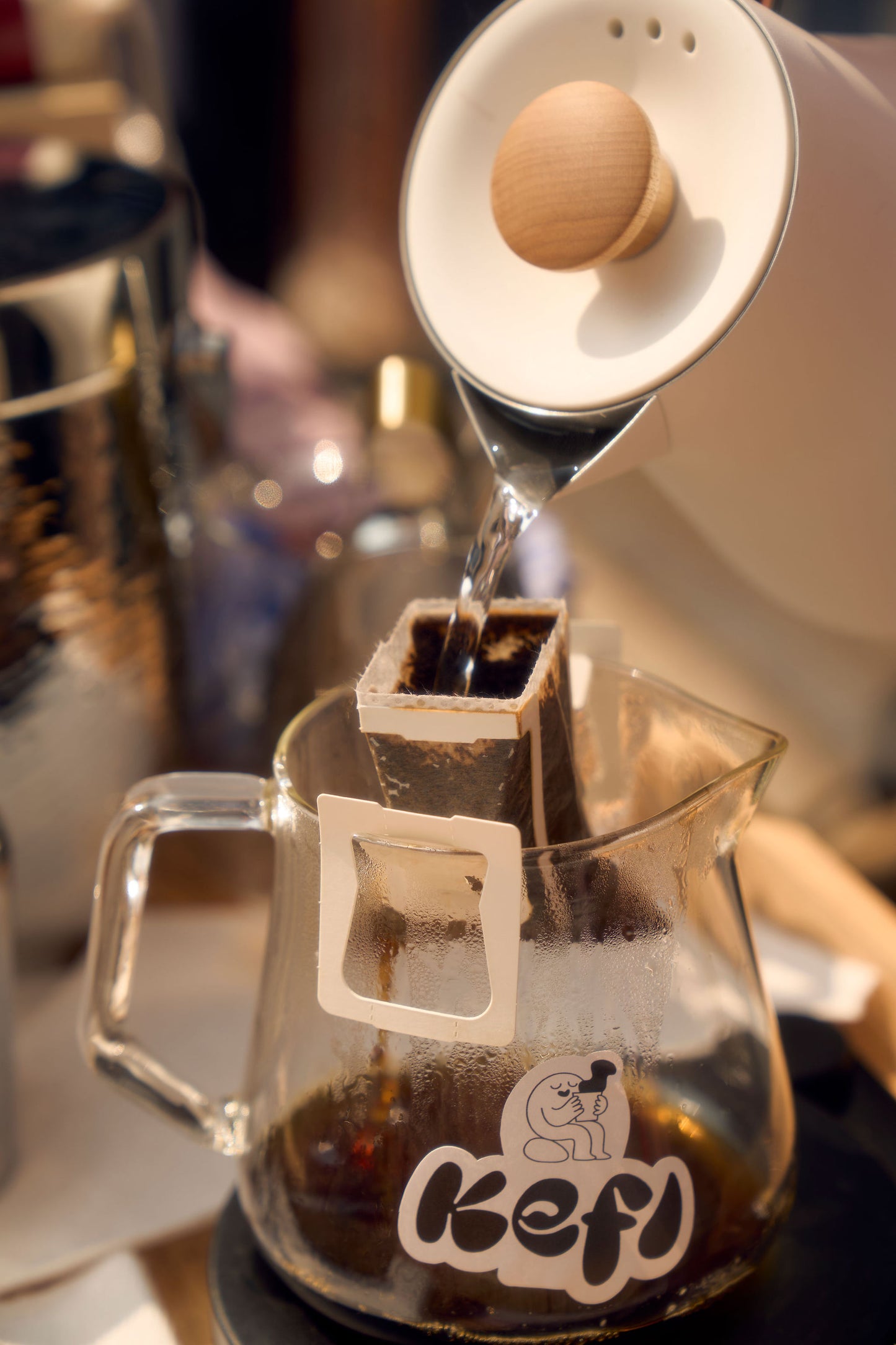 Hot water is being poured from a white kettle into a Kefi Coffee single-serve pour-over filter positioned over a glass carafe. The carafe features the Kefi Coffee logo, depicting a joyful character sipping from a cup. The scene is cozy, with coffee brewing equipment and a soft background, emphasizing the brewing process and rich coffee being extracted into the carafe.