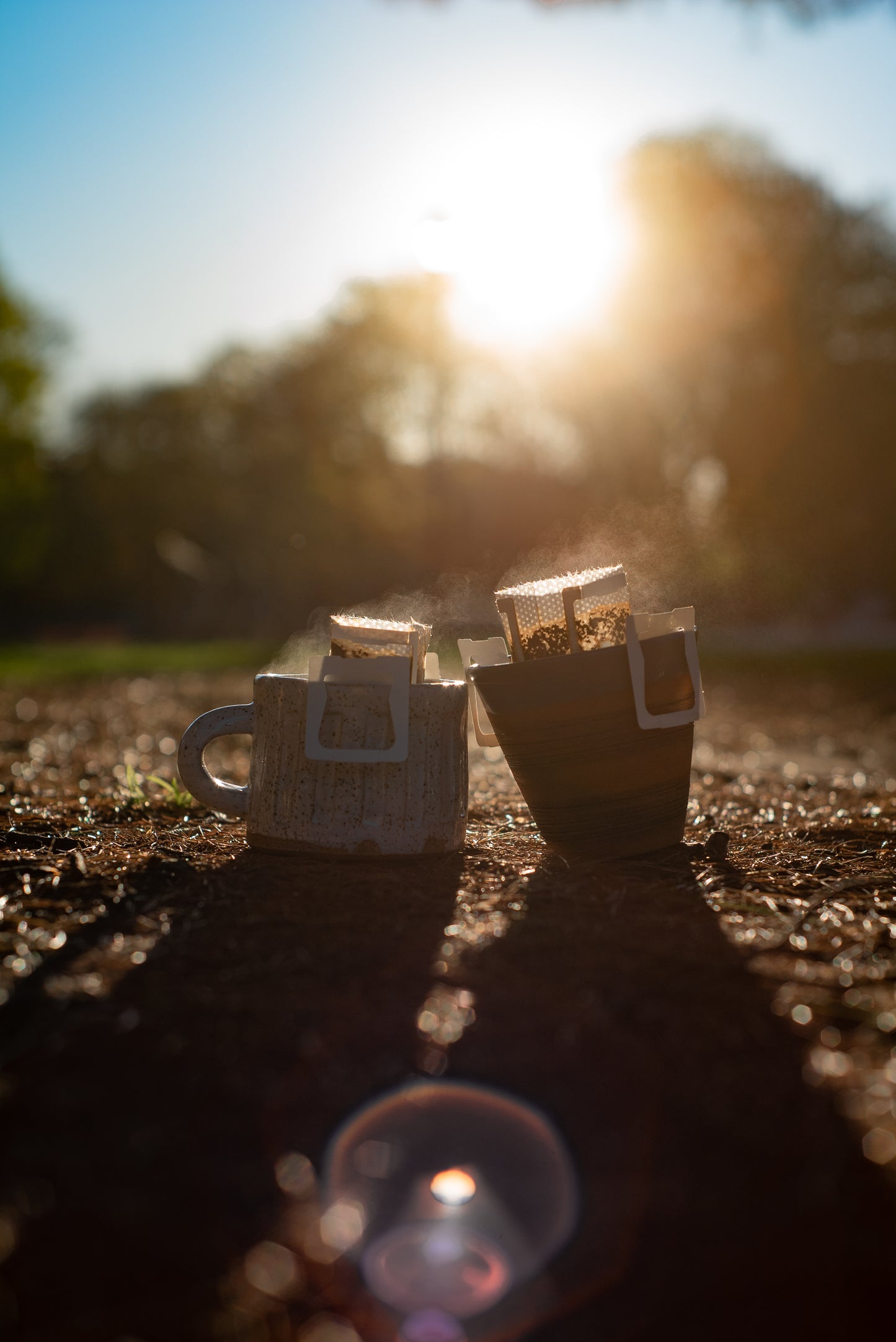 Two mugs with Kefi Coffee pour-over coffee filters are placed on the ground outdoors. Steam is rising from the filters as the sun sets in the background, casting a warm, golden light over the scene. The left mug is white with a textured design, and the right mug has a striped pattern. The background is slightly blurred with trees and natural surroundings, creating a peaceful and serene atmosphere.