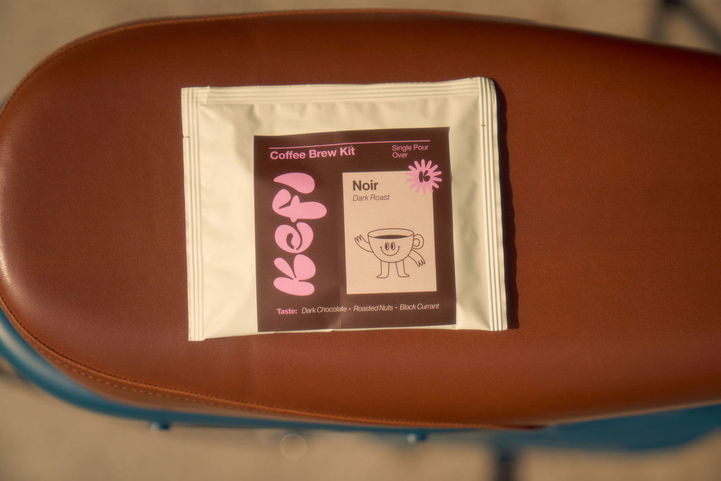 A Kefi Coffee “Noir Dark Roast” single-serve pour-over coffee kit placed on a brown leather surface. The packaging features Kefi’s logo in soft pink letters, alongside a playful illustration of a smiling coffee cup with arms and legs. The label indicates tasting notes of dark chocolate, roasted nuts, and black currant.