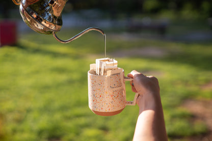 A hand holding a white speckled mug with colorful dots outdoors. The mug has a Kefi Coffee pour-over coffee filter attached, and a stream of water is being poured from a gooseneck kettle into the filter. The background shows a blurred grassy area with sunlight creating a warm and inviting atmosphere, like in the park or picnic.