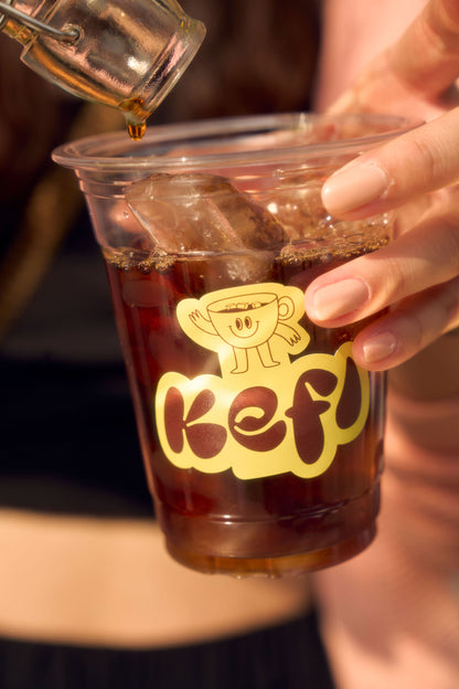 A close-up of iced coffee being poured into a clear plastic cup branded with the Kefi Coffee logo. The logo features a cheerful coffee cup character, and the person holding the cup has manicured nails. Ice cubes float at the top of the dark, freshly brewed coffee as it fills the cup, with a soft, blurred background adding a warm ambiance.