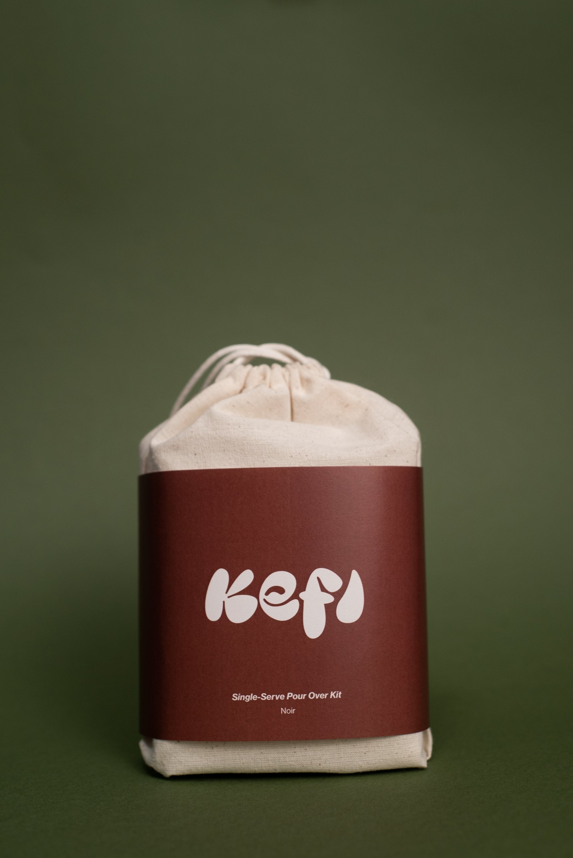 A beige cotton drawstring bag with a maroon label featuring the word ‘Kefi’ in white, bold, playful font. Below the brand name, the text reads ‘Single-Serve Pour Over Kit’ and ‘Noir’. The bag is set against a muted green background, highlighting its minimalist and clean design.