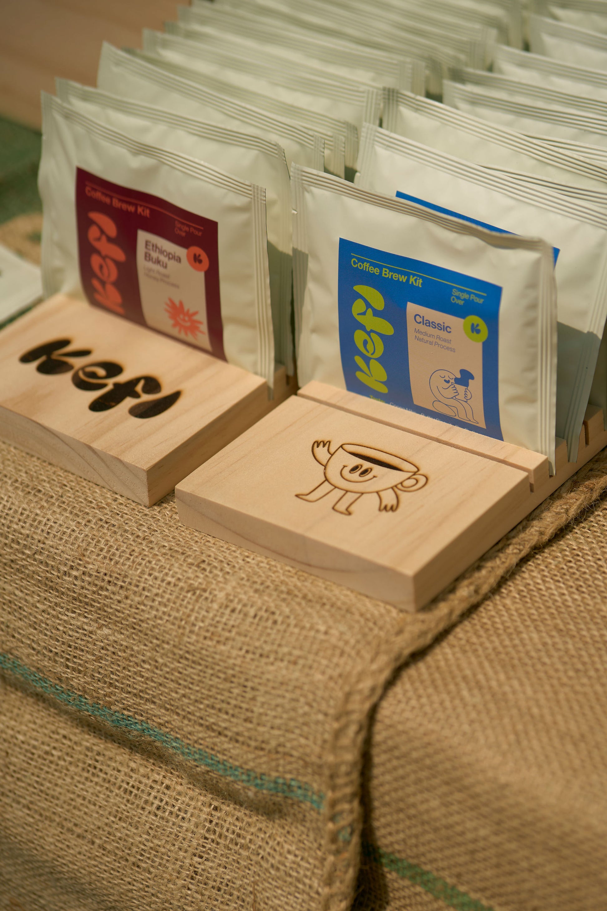 A display of Kefi Coffee single-serve pour-over kits neatly arranged in wooden holders on a burlap-covered table. The kits are presented in two variations: the “Ethiopia Buku” with a red and orange label, and the “Classic” blend with a blue and green label. The wooden holders feature the Kefi Coffee logo and a playful coffee cup character. The setup has a rustic, artisanal feel, with the burlap adding texture and warmth to the display.