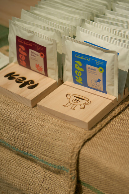 A display of Kefi Coffee single-serve pour-over kits neatly arranged in wooden holders on a burlap-covered table. The kits are presented in two variations: the “Ethiopia Buku” with a red and orange label, and the “Classic” blend with a blue and green label. The wooden holders feature the Kefi Coffee logo and a playful coffee cup character. The setup has a rustic, artisanal feel, with the burlap adding texture and warmth to the display.