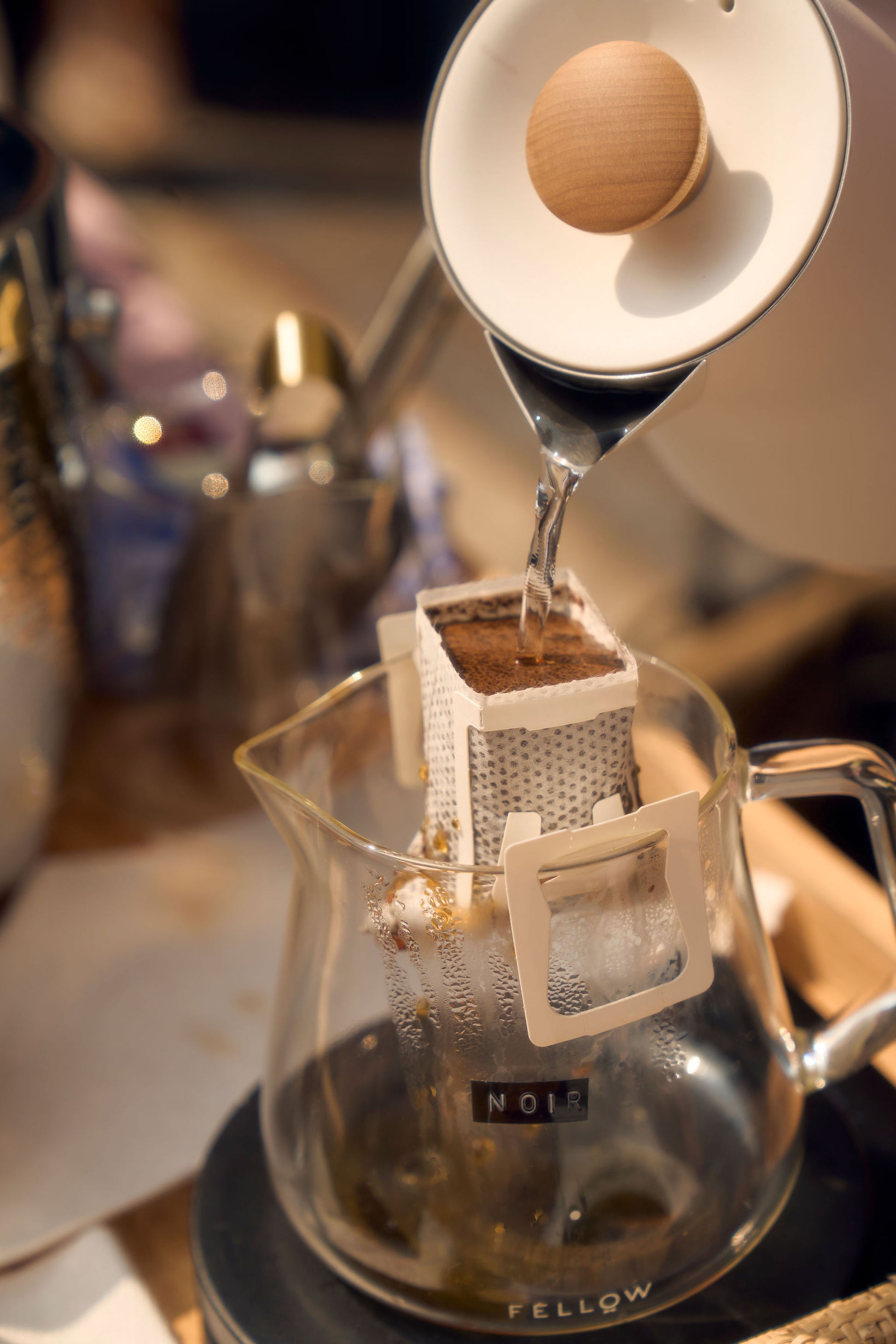 A close-up shot of hot water being poured from a kettle into a single-serve pour-over coffee filter positioned over a glass carafe. The filter, containing coffee grounds, is part of Kefi Coffee’s brew kit, and the glass carafe has a label marked “Noir.” The scene is set on a wooden table with a cozy, warm ambiance, and brewing equipment visible in the background