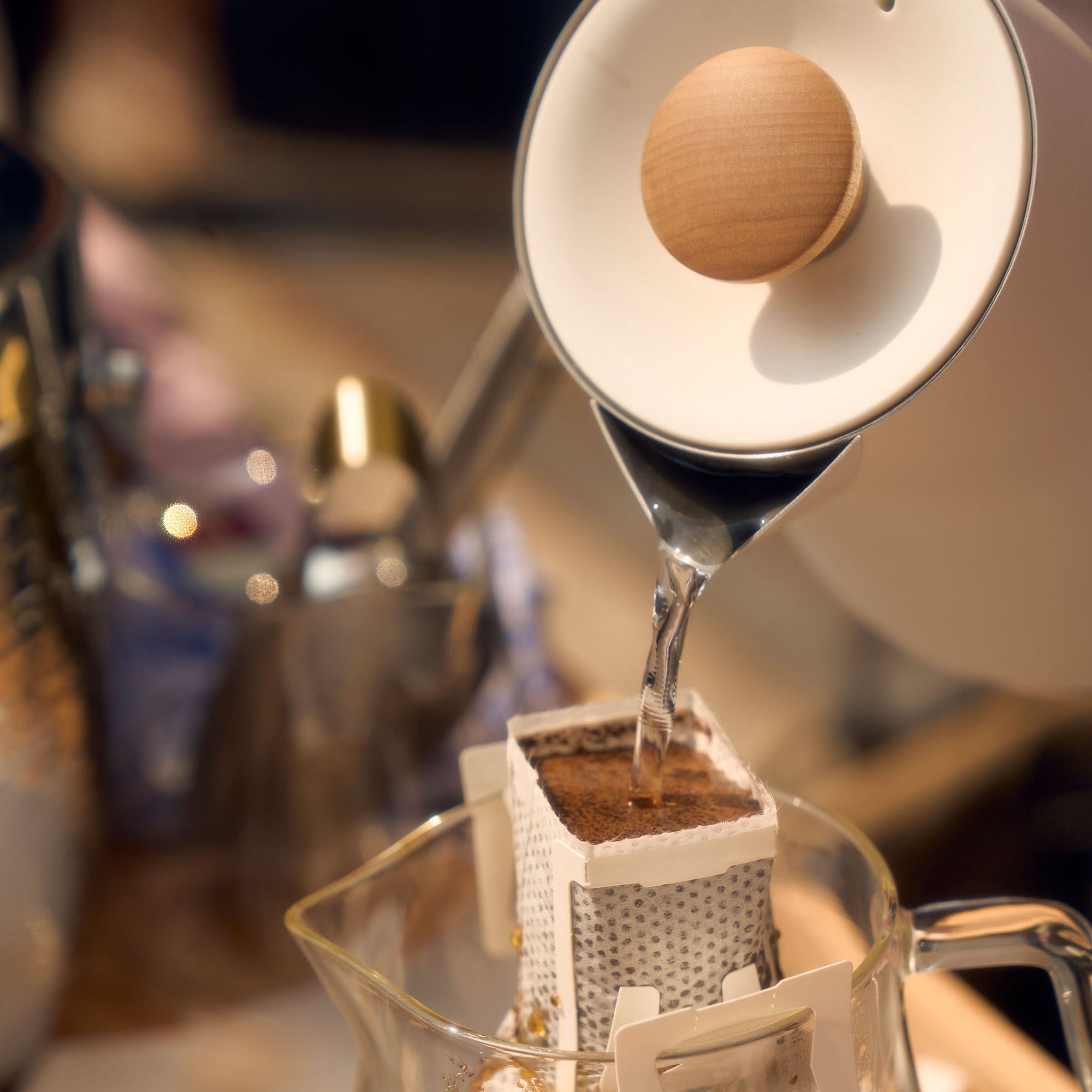 A close-up shot of hot water being poured from a kettle into a single-serve pour-over coffee filter positioned over a glass carafe. The filter, containing coffee grounds, is part of Kefi Coffee’s brew kit, and the glass carafe has a label marked “Noir.” The scene is set on a wooden table with a cozy, warm ambiance, and brewing equipment visible in the background