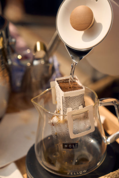 A close-up shot of hot water being poured from a kettle into a single-serve pour-over coffee filter positioned over a glass carafe. The filter, containing coffee grounds, is part of Kefi Coffee’s brew kit, and the glass carafe has a label marked “Noir.” The scene is set on a wooden table with a cozy, warm ambiance, and brewing equipment visible in the background