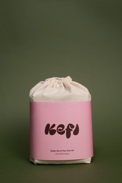 A beige cotton drawstring bag with a pink label featuring the word ‘Kefi’ in white, bold, playful font. Below the brand name, the text reads ‘Single-Serve Pour Over Kit’ and ‘Noir’. The bag is set against a bright yellow background, highlighting its minimalist and clean design.