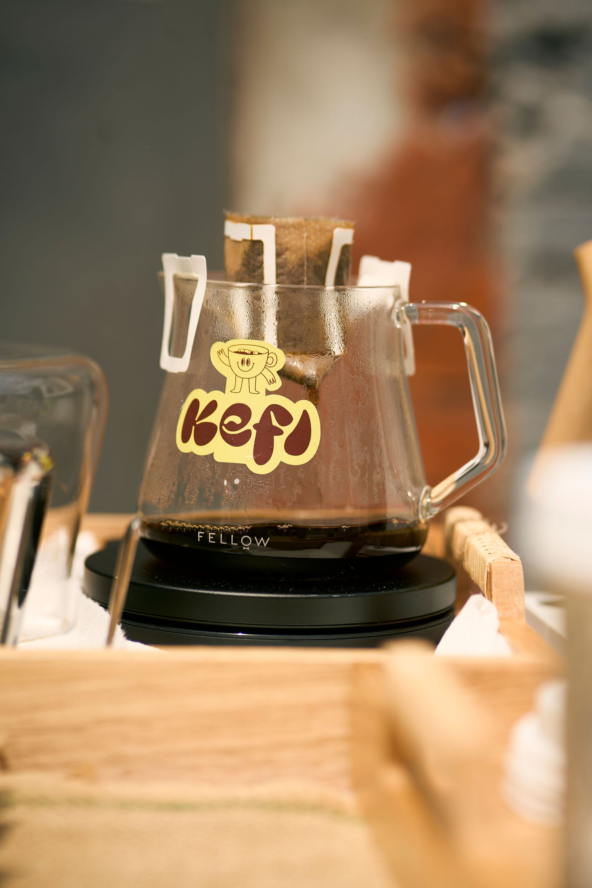 A glass carafe with the Kefi Coffee logo, showing a cheerful character, sits on a wooden surface while brewing coffee. The single-serve pour-over filter is suspended above the carafe as fresh coffee drips into the container. The scene is warm and inviting, with the carafe resting on a scale, emphasizing the precision of the brewing process. The soft lighting highlights the rich coffee as it collects in the carafe.