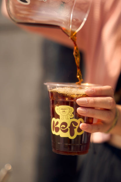 A hand holds a clear plastic cup with the Kefi Coffee logo as freshly brewed coffee is poured into it from a glass carafe. The coffee streams into the cup, creating a rich, dark layer of liquid. The yellow Kefi logo, featuring a joyful character, is prominently displayed on the cup. The person pouring wears a soft pink top, and the background is softly blurred.