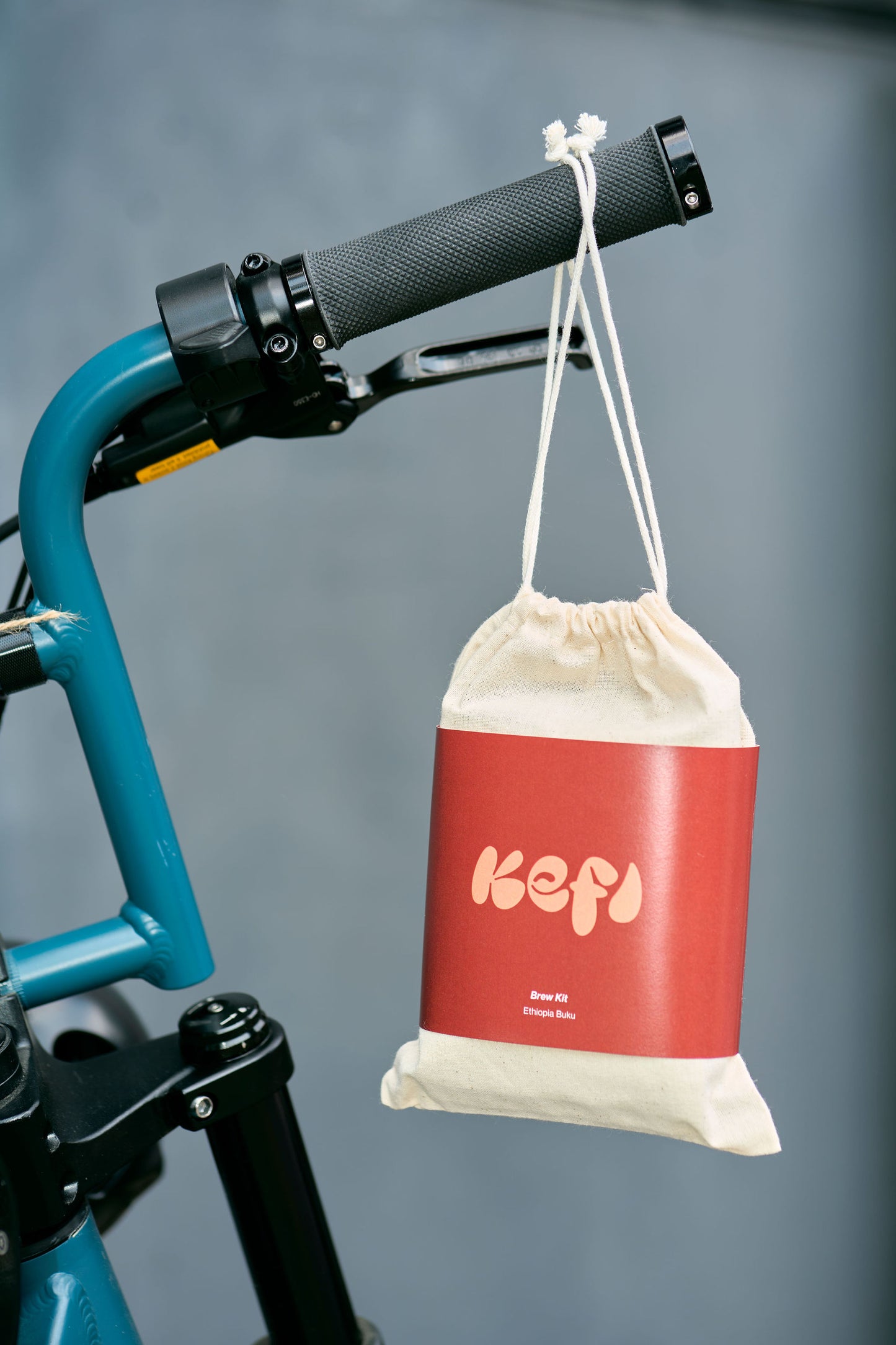 A reusable cotton drawstring bag with a red Kefi Coffee label hangs from the handlebar of a teal-colored bicycle. The red label features the Kefi logo in soft pink lettering. The minimalist design of the packaging complements the sleek and modern look of the bike. The bag is positioned against a neutral, blurred background, emphasizing the product and its portability, ideal for coffee lovers on the go.