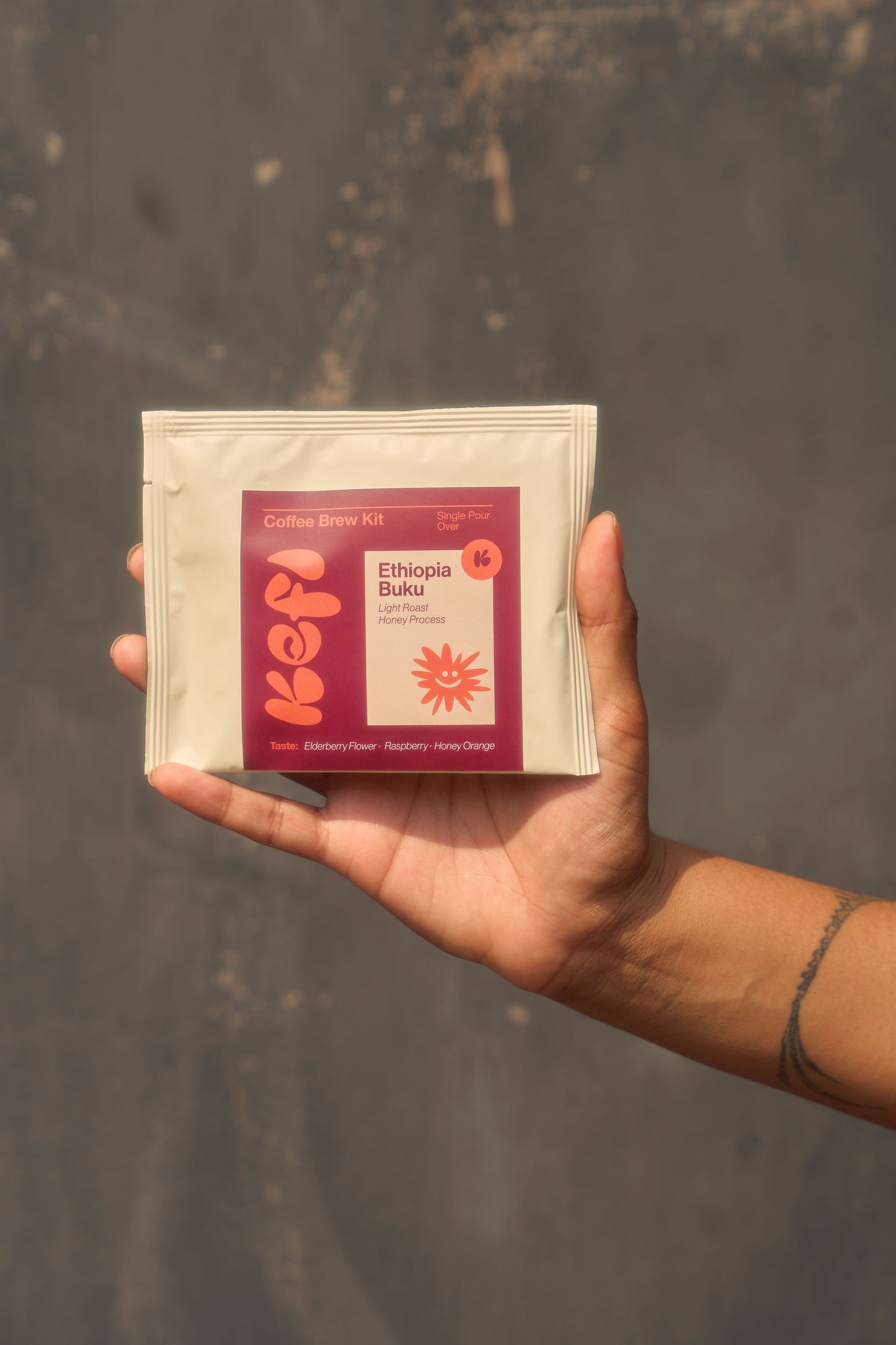 A hand holds a Kefi Coffee “Ethiopia Buku” a coffee brew kit against a neutral, textured background. The packaging features a vibrant design with a red and orange label displaying the Kefi logo and a sunburst graphic. The tasting notes on the package describe the coffee as offering a blueberry, honey, and blood orange flavor profile. The person holding the kit has a minimalist tattoo on their forearm, and the focus is on the product, framed by the natural, warm lighting.