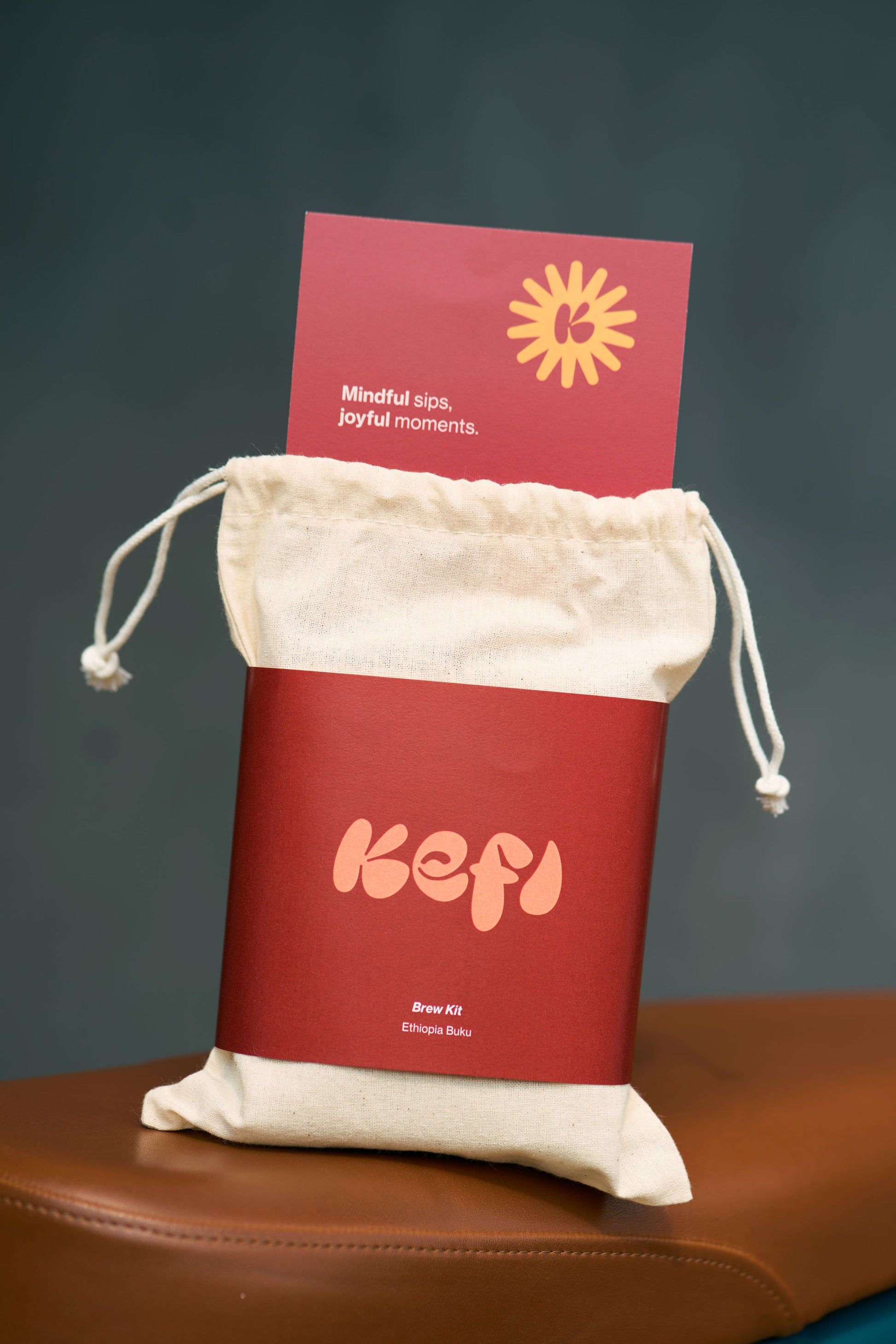 A Kefi Coffee brew kit package displayed in a simple, reusable cotton drawstring bag. The bag features a sleek red label with the Kefi logo in soft pink lettering. A card is tucked into the top of the bag, reading “Mindful sips, joyful moments” with a sunburst graphic above it. The bag rests on a brown leather surface, and the background is a soft, neutral tone, emphasizing the minimalist and sustainable packaging design.