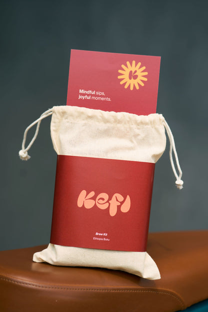 A Kefi Coffee brew kit package displayed in a simple, reusable cotton drawstring bag. The bag features a sleek red label with the Kefi logo in soft pink lettering. A card is tucked into the top of the bag, reading “Mindful sips, joyful moments” with a sunburst graphic above it. The bag rests on a brown leather surface, and the background is a soft, neutral tone, emphasizing the minimalist and sustainable packaging design.