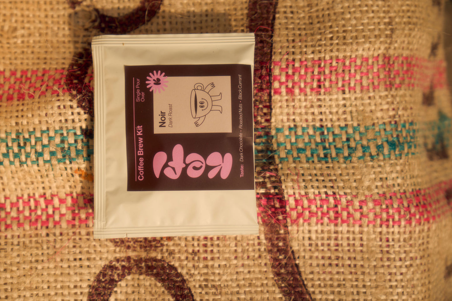 “Noir Dark Roast” coffee pour-over brew kit rests on a textured burlap surface with colorful woven patterns in red, green, and pink. The packaging features a dark brown label with soft pink Kefi branding and an illustration of a smiling coffee cup character. Tasting notes of dark chocolate, roasted nuts, and black currant are listed on the bottom of the package, highlighting the rich flavors of the dark roast. The natural burlap background adds a rustic and earthy feel to the presentation.