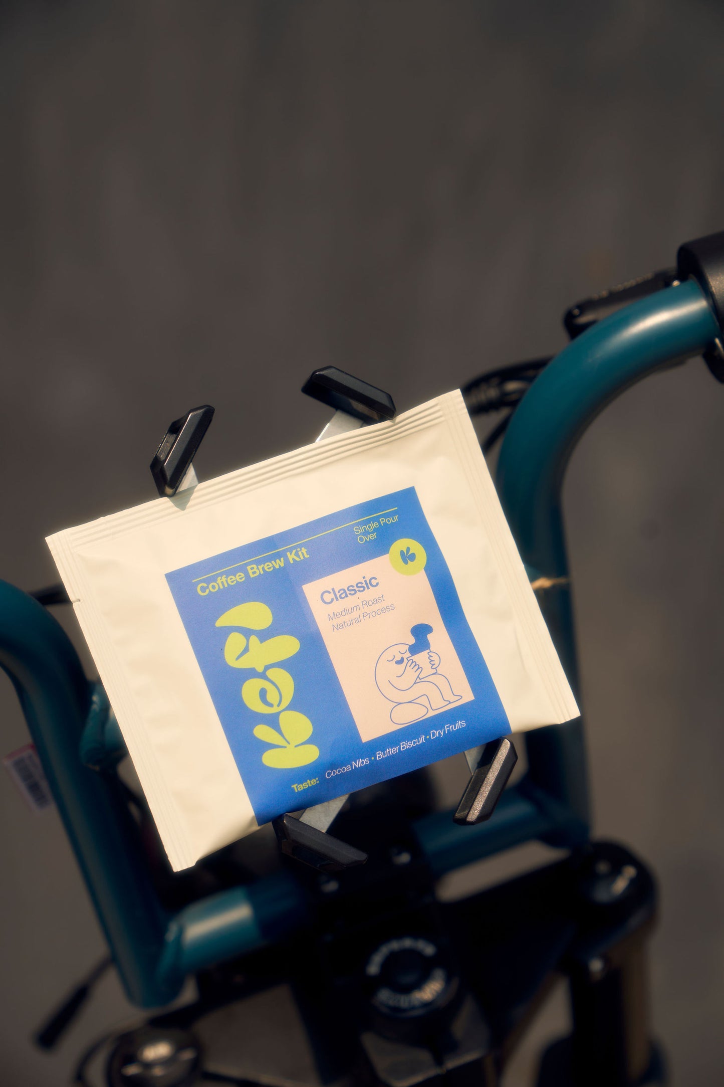 Classic” single-serve pour-over coffee kit resting on the handlebars of a teal-colored bicycle. The packaging features a bright blue and yellow label with the Kefi logo and an illustration of a person enjoying a cup of coffee. The background is softly blurred, emphasizing the coffee kit and the bike handlebars. The label highlights tasting notes of smooth and balanced coffee flavors.