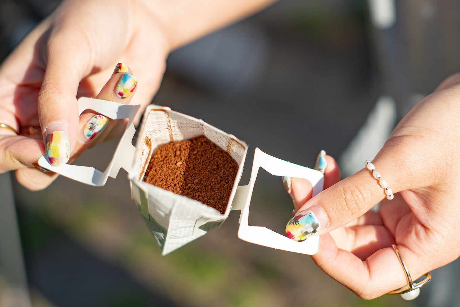 A close-up of a person holding a Kefi Coffee pour-over coffee filter filled with ground coffee, ready to brew. The person’s nails are painted with a colorful abstract design, and they are wearing a pearl ring on one finger. The filter is being held open, showing the coffee grounds inside, with an outdoor background blurred in the distance.