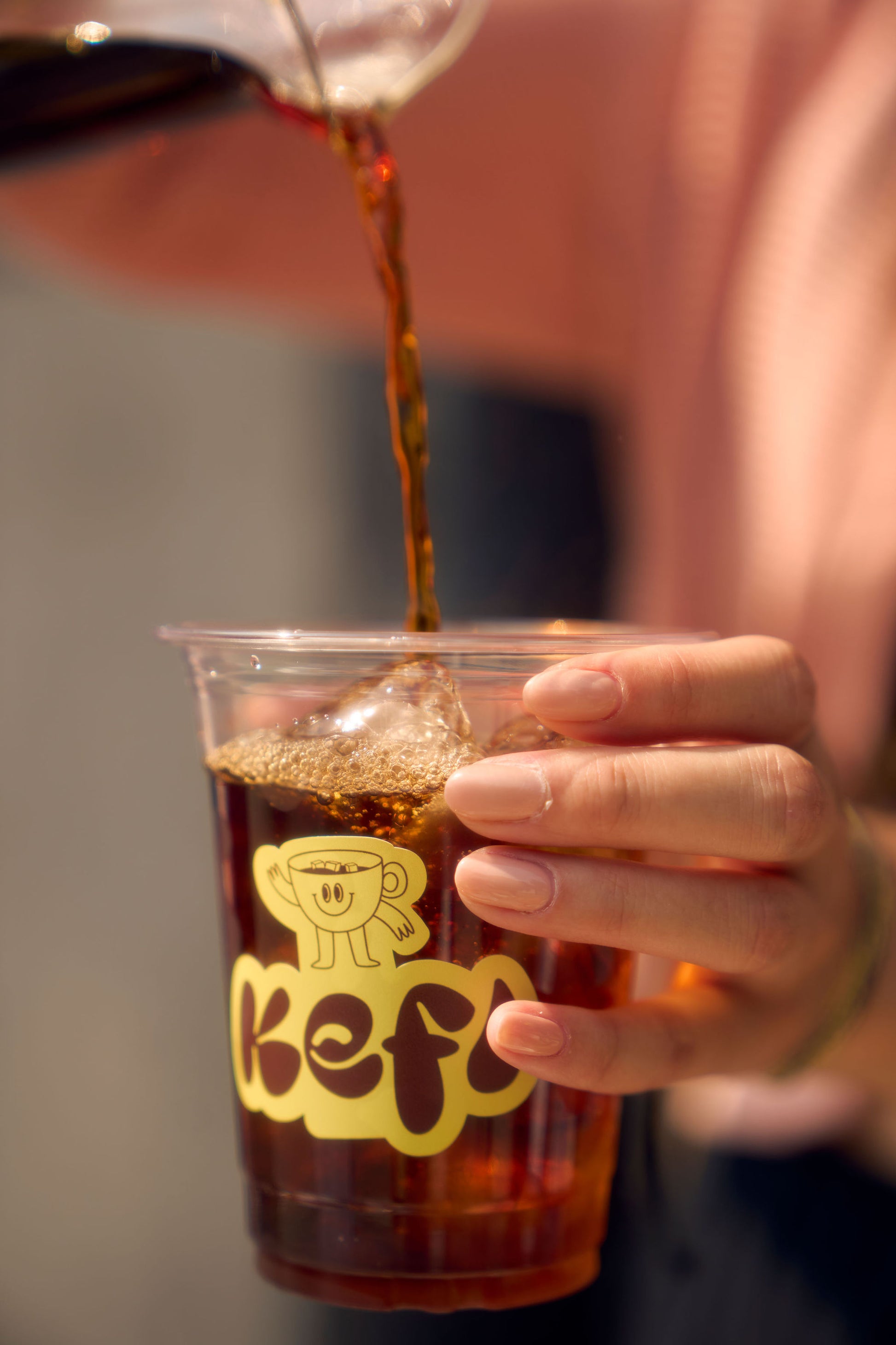 A close-up of a clear plastic cup with the Kefi Coffee logo, featuring a smiling coffee cup character, as freshly brewed coffee is poured into it from a glass carafe. The person holding the cup has neatly manicured nails and is gently tilting the cup to allow the coffee to flow in, creating a rich dark layer of liquid. The background is softly blurred, bringing attention to the vibrant coffee and the branding on the cup.