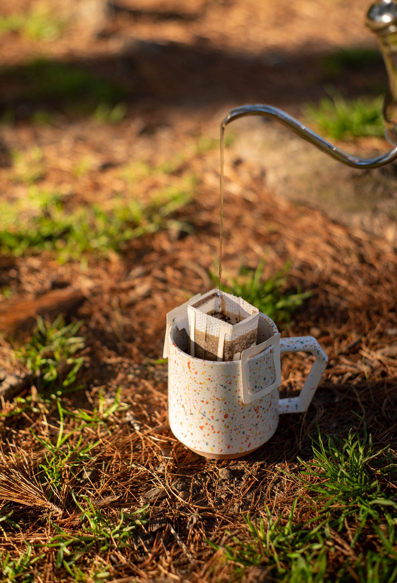 A white speckled mug with colorful dots is placed on the ground outdoors. The mug has a pour-over coffee filter attached, and a stream of hot water is being poured from a gooseneck kettle into the filter. The ground is covered with pine needles and patches of grass, creating a natural and rustic setting.