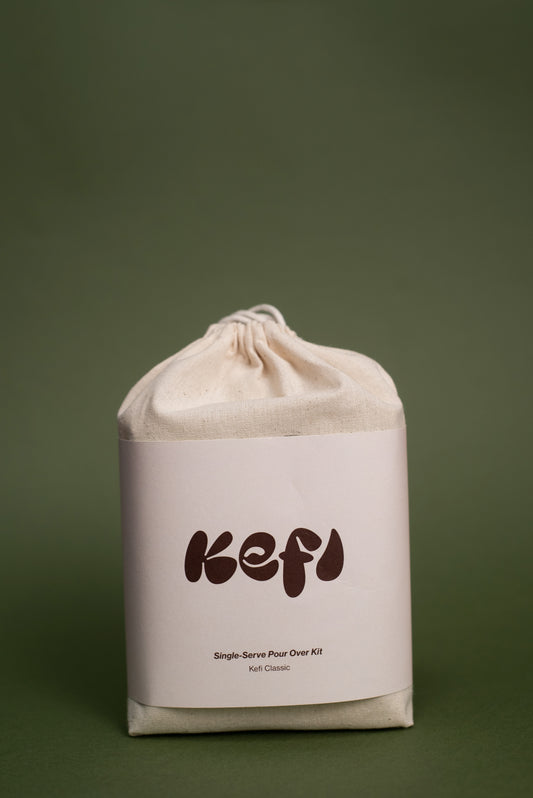 A beige cotton drawstring bag with a light beige label featuring the word ‘Kefi’ in dark brown, bold, playful font. Below the brand name, the text reads ‘Single-Serve Pour Over Kit’ and ‘Kefi Classic’. The bag is set against a muted green background, highlighting its minimalist and clean design