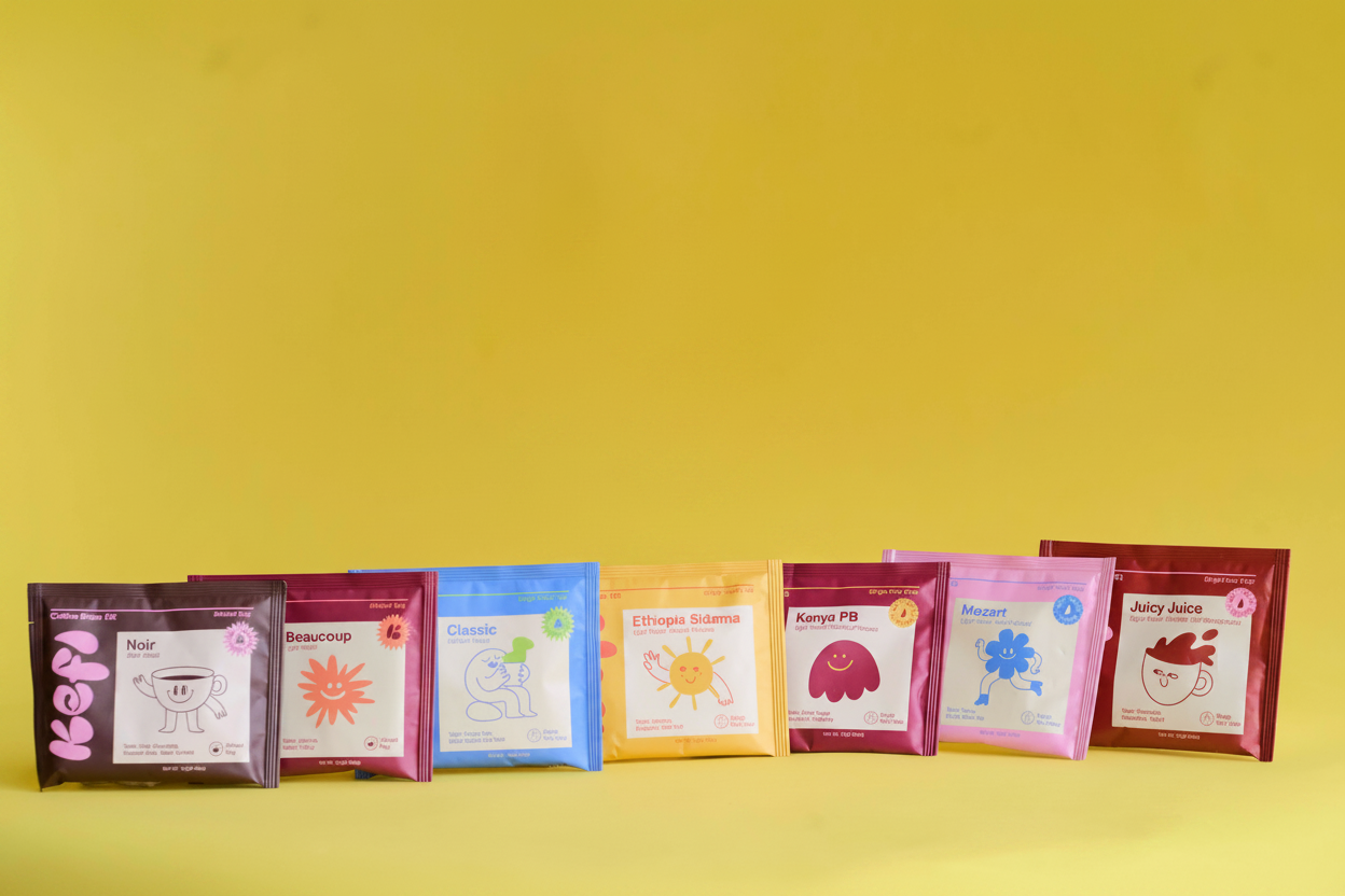 Colorful lineup of Kefi Coffee single-serve pour-over brew kits arranged in front of a drawstring cotton bag with a pink Kefi label. Each coffee pouch features a playful character design and bright color, representing different roast profiles and origins such as Noir, Beaucoup, Classic, Ethiopia Sidama, Kenya PB, Mozart, and Juicy Juice. Set against a solid yellow background